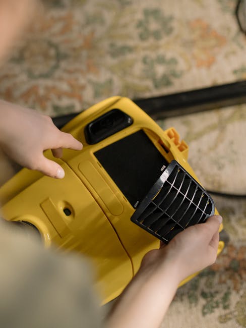 Close-up view of a person's hands operating a yellow portable air mover or blower used for surface cleaning or rapid drying in a room with a patterned carpet and soft natural lighting. The device feat