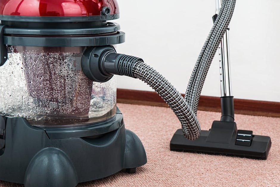 A person kneeling on a patterned carpet in a residential room, using a yellow professional steam cleaner with a black hose attachment for surface cleaning. The individual is wearing a beige jacket and